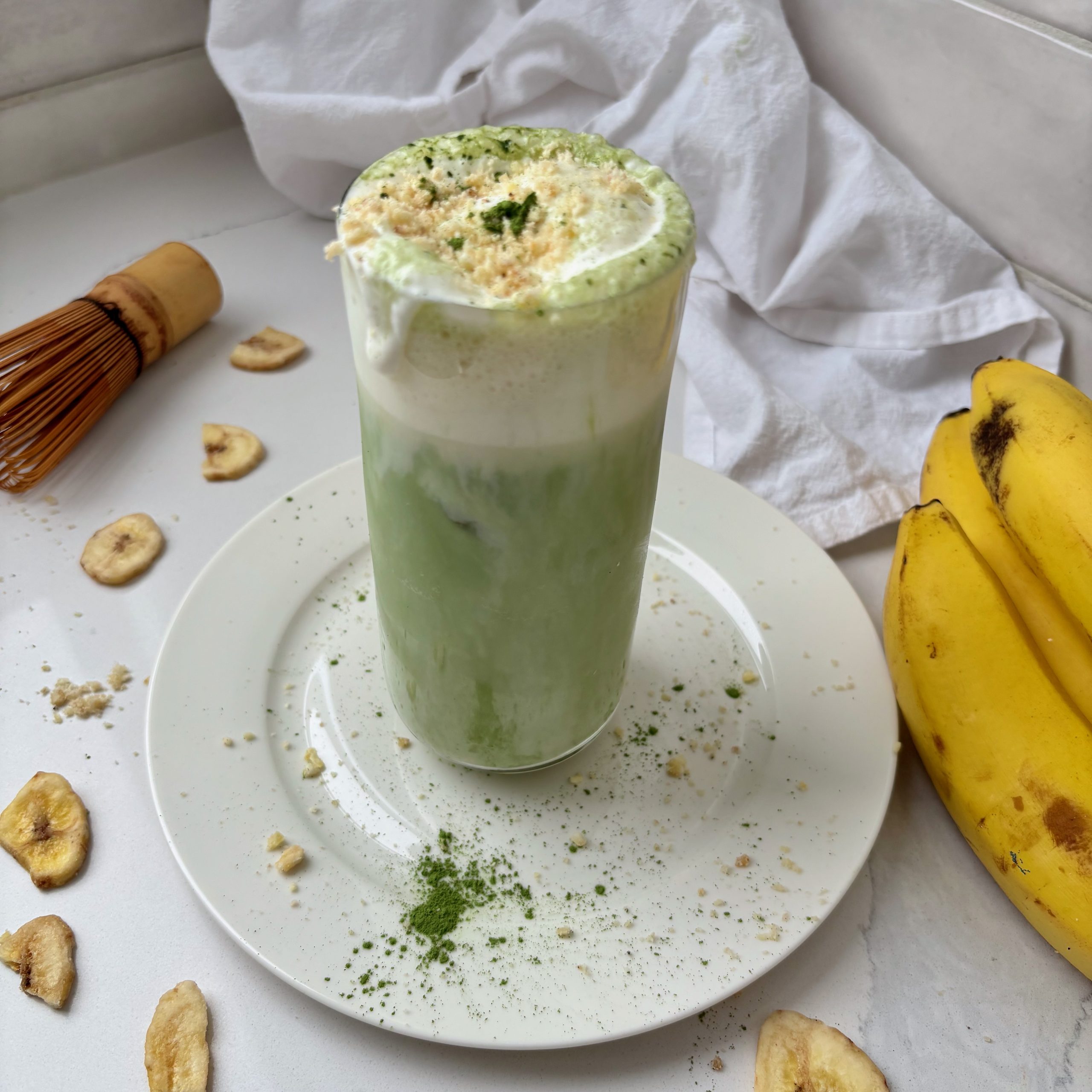 Banana Cream Matcha Latte in glass with bananas and matcha whisk