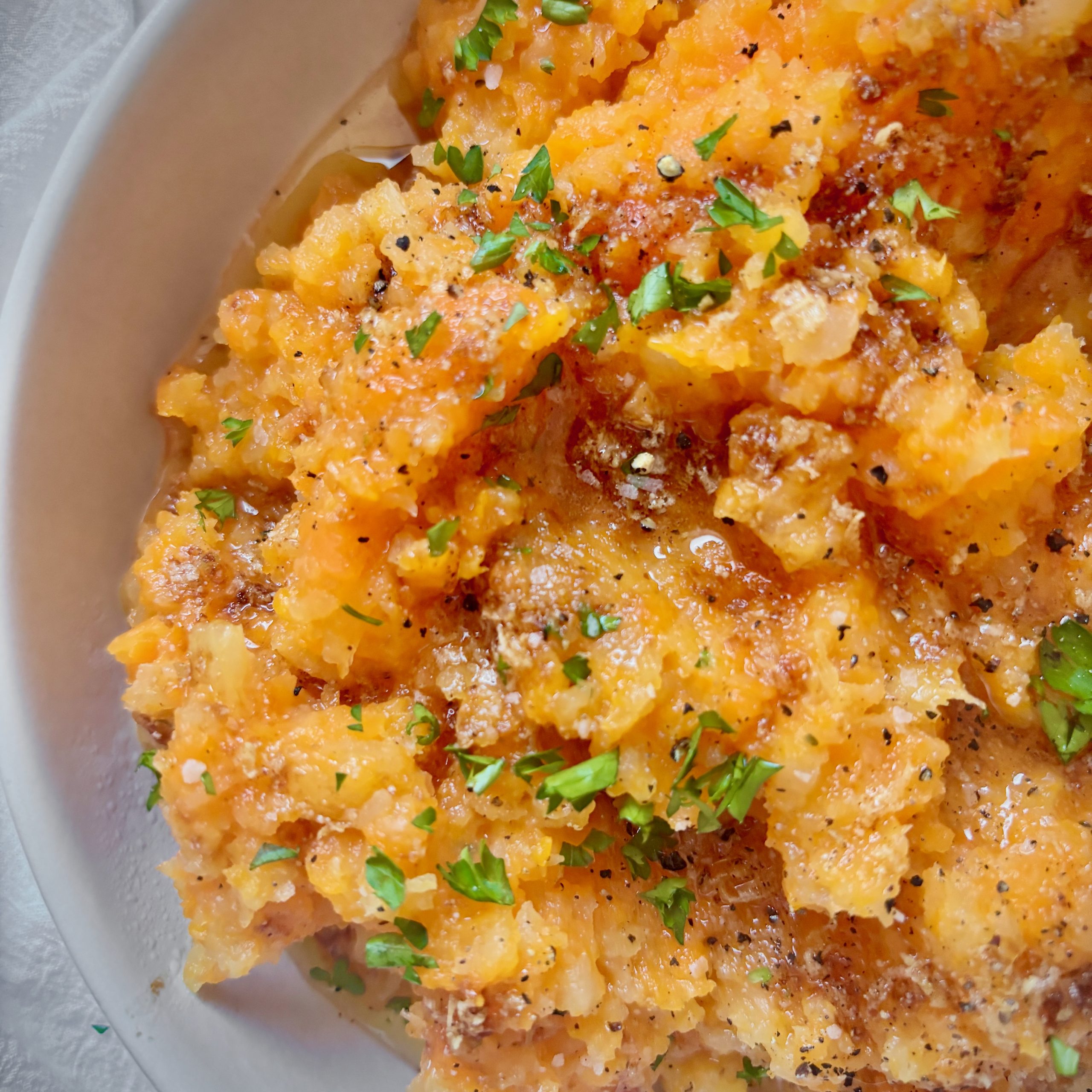 A bowl of mashed carrots and rutabaga topped with garlic brown butter and chopped parsley, showing a rustic, slightly chunky texture.