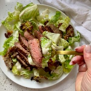 A hand holding a gold fork with a bite of caesar salad with steak and a crispy garlic parmesan smashed potato crouton.