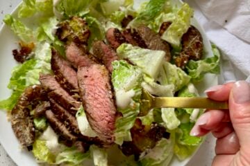 A hand holding a gold fork with a bite of caesar salad with steak and a crispy garlic parmesan smashed potato crouton.