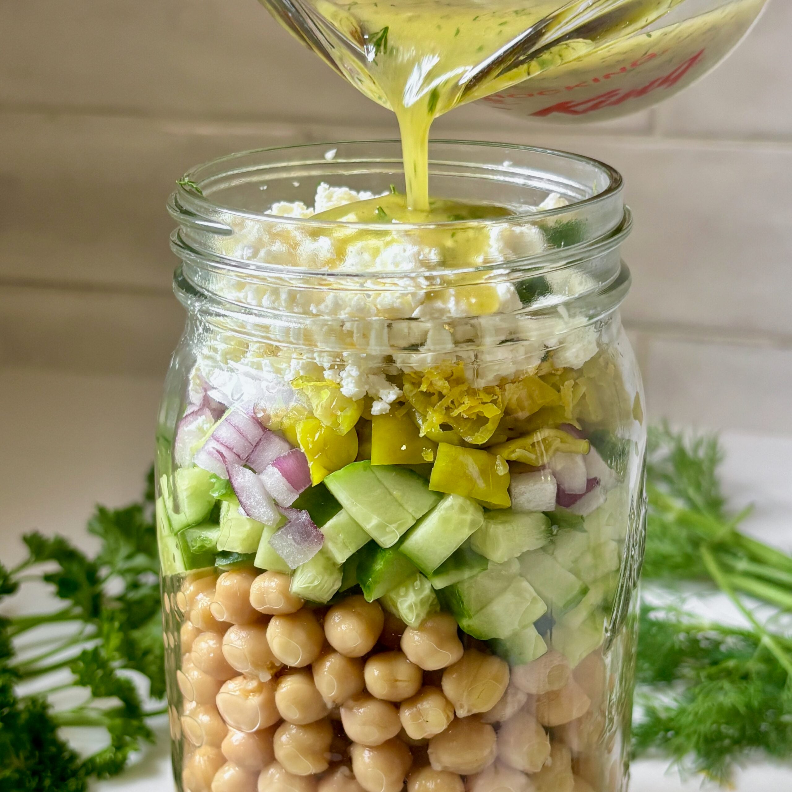 Pouring the lemon-dill dressing over chickpeas and feta in a wide-mouth mason jar.