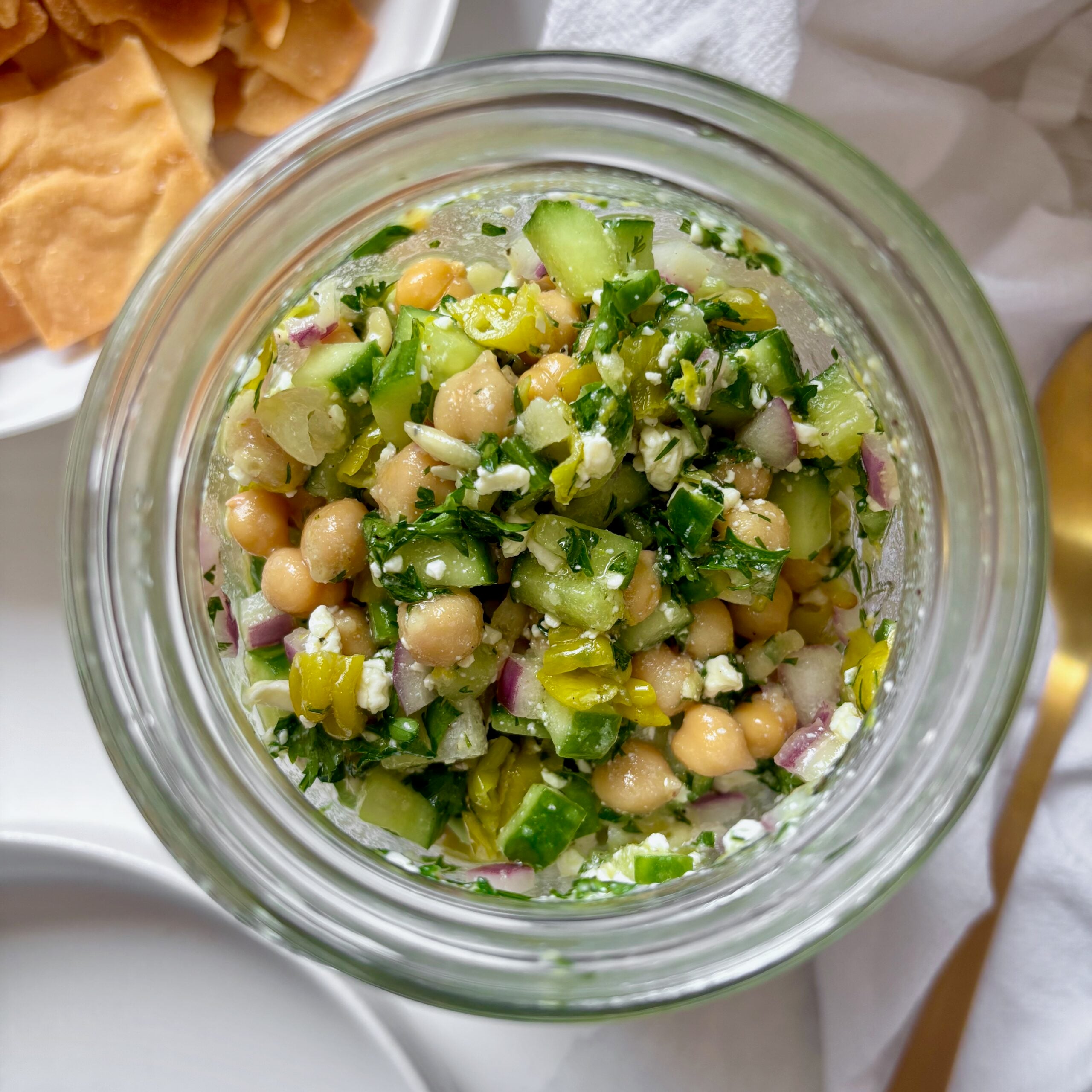 A glass jar filled with chickpeas, feta, and cucumbers, topped with fresh dill and lemon zest.
