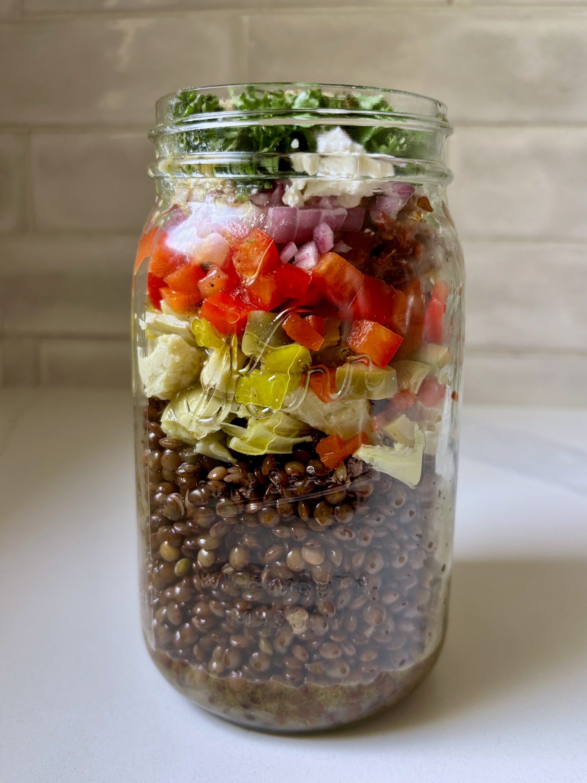 A large glass mason jar filled with layered Mediterranean marinated lentil salad featuring distinct layers of cooked lentils, chopped artichokes, bright red peppers, and crumbled feta cheese